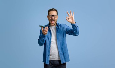 İşadamı sevinçle bağırıyor ve mavi arka planda cep telefonu kullanırken tamam işareti gösteriyor.