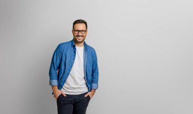 Elleri ceplerinde, gülümseyen ve beyaz arka planda tek başına duran kendinden emin genç bir iş adamı.