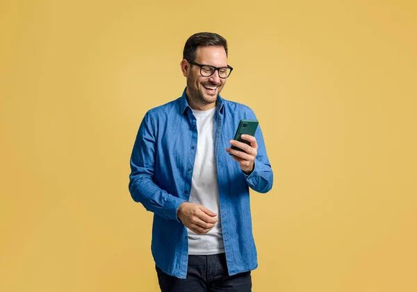 Happy handsome businessman online messaging over smart phone while standing on yellow background