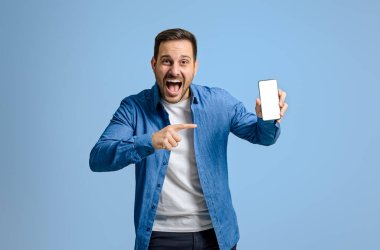 Heyecanlı erkek girişimci çığlık atıyor ve mavi arkaplan üzerindeki boş akıllı telefon ekranına işaret ediyor