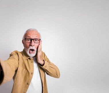 Endişeli kıdemli işadamı yüzüne dokunuyor ve beyaz arka planda şok halinde kameraya bakıyor.