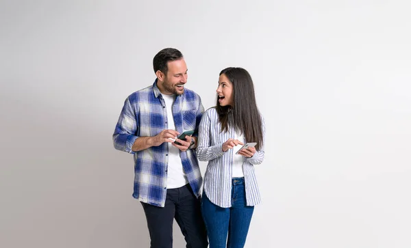 Cheerful young couple with mobile phones looking at each other excitedly against white background