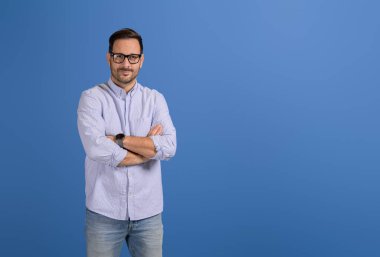 Kolları çapraz ve gözlükleri mavi arka planda kameraya bakan kendinden emin bir erkek profesyonel.