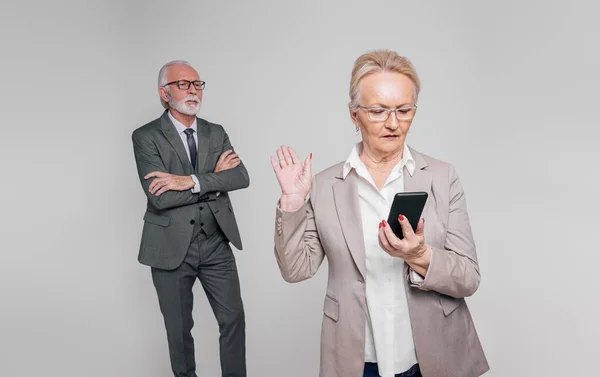 Old businesswoman checking mobile phone and showing stop gesture to businessman on white background