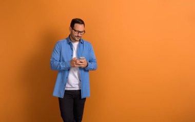 Gülümseyen yakışıklı işadamı sosyal medya uygulamalarını izole edilmiş turuncu arka planda cep telefonundan kullanıyor.