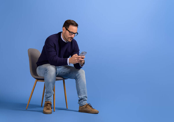 Focused serious businessman checking social media apps over smart phone on chair on blue background