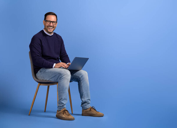 Portrait of cheerful male manager marketing online over computer on chair against blue background