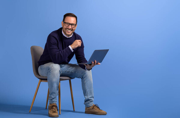 Successful male manager with laptop smiling at camera while sitting on chair over blue background