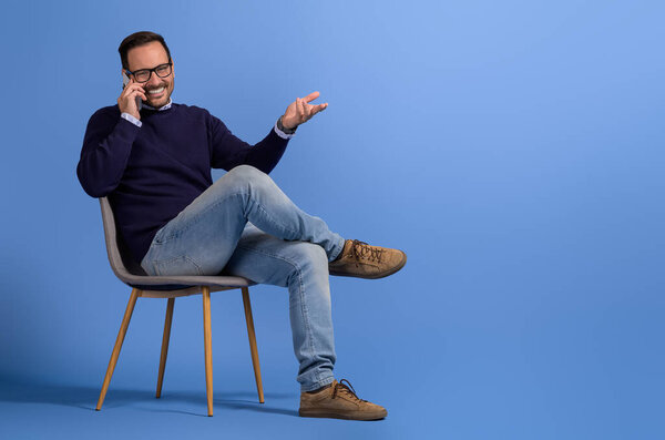 Happy businessman discussing ideas over mobile phone while sitting on chair against blue background