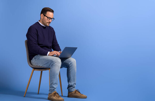 Portrait of male entrepreneur reading e-mails over laptop and sitting on chair over blue background