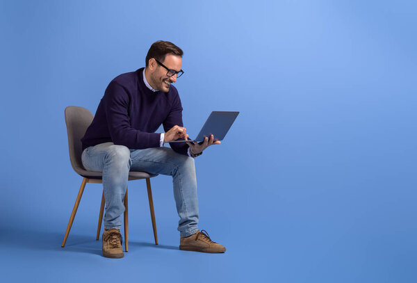 Smiling businessman checking online websites over laptop while sitting on chair over blue background