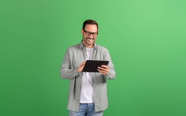 Gözlüklü gülümseyen genç işadamı yeşil arka planda dijital tablet üzerinden e-postaları okuyor.