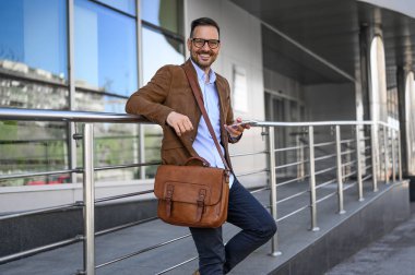 Yakışıklı girişimci portresi cep telefonu kullanıyor ve şehrin dışında kameraya gülümsüyor