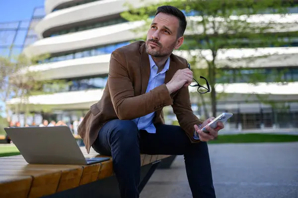 Elgondolkodó Fiatal Üzletember Mobiltelefonnal Laptoppal Félrenéz Miközben Padon Városban Stock Kép