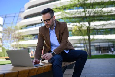 Ciddi bir erkek çalışanın modern şehirde bankta otururken kablosuz bilgisayardan raporunu analiz etmesi
