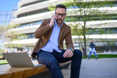 Modern şehirdeki bankta kablosuz bilgisayarda çalışırken telefonda konuşan yakışıklı bir adam.