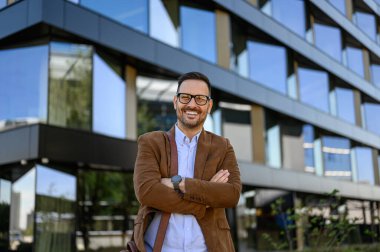 Gözlüklü iyimser yöneticinin portresi ve kolları çaprazlanmış modern binaya karşı duruyor.