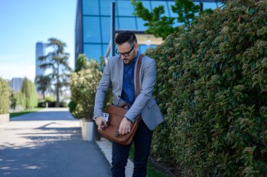 Gözlüklü yakışıklı yönetici şehir sokaklarında bitkilerin yanında dururken kahverengi laptop çantası takıyor.