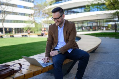 Gözlüklü olumlu işadamı, şehirdeki ahşap bankta otururken laptop üzerinden veri analiz ediyor.