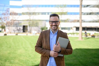 Kablosuz bilgisayarla çimlere ve binaya gülümseyen kendinden emin iş adamının portresi