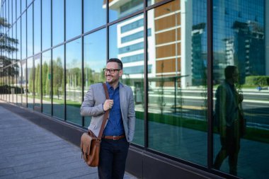 Çantasıyla başarılı bir banliyö çalışanı, modern ofis binasının yanındaki kaldırıma bakıyor ve dikiliyor.