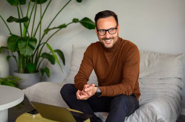 Ofisteki kanepede dizüstü bilgisayar başında çalışırken gözlüklü mutlu bir işadamı dikkatle başka tarafa bakıyor.