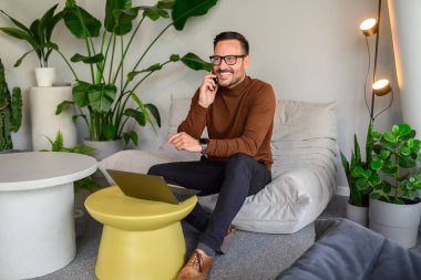 Gülümseyen işadamı proje detaylarını telefonda konuşuyor ve rahat koltukta dizüstü bilgisayar kullanıyor.