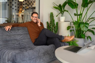 Ofisteki koltukta dinlenirken gözlüklü, cep telefonuyla konuşan pozitif erkek girişimci.