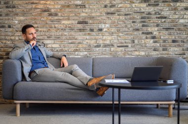 Ciddi bir finansal danışman gözlük tutuyor ve ofisteki kanepeye bakarken düşünüyor.