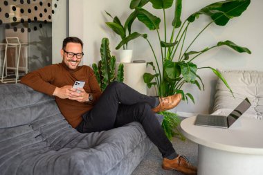 Modern ofisteki dizüstü bilgisayarla koltukta dinlenirken akıllı telefondan mesajlaşan orta büyüklükte bir profesyonel.