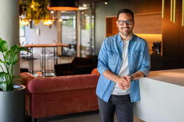 Yakışıklı mühendisin portresi, gözlüklü ve elleri bağlı gülümseyen ve modern ofiste ayakta duran.