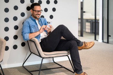 Ofis koltuğunda otururken sosyal medya uygulamalarını cep telefonundan kontrol eden gözlüklü bir iş adamı.