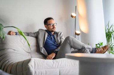 Gözlüklü, düşünceli bir erkek proje yöneticisi gözlerini kaçırıyor ve modern ofiste koltukta dinleniyor.