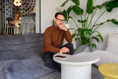 Ofisteki kanepede otururken dizüstü bilgisayarı inceleyen eli çenesinde dalgın bir erkek muhasebeci.