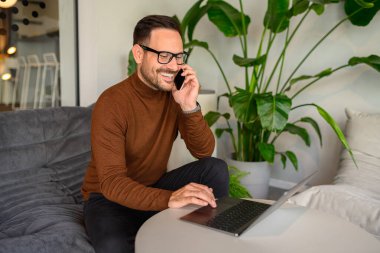 Neşeli orta yaşlı bir adam iş görüşmeleri yapıyor ve modern ofiste dizüstü bilgisayarla çalışıyor.