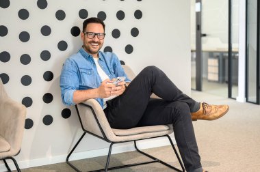 Gözlüklü genç adam modern ofiste koltukta dinlenirken akıllı telefondan mesaj atıyor.