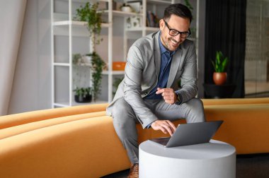 Ofiste sarı koltukta otururken dizüstü bilgisayarından gülümseyen ve e-postaları okuyan gözlüklü yakışıklı CEO.