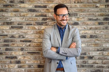 İyimser iş adamının portresi. Kollarını kavuşturup düşüncelerinin üzerinden gülümsüyor. Ofiste tuğla duvar var.