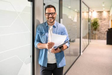 Bilgisayarı ve belgesi olan özgüvenli erkek programcının portresi modern ofiste cam duvarın yanında duruyor.