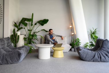 Ofisteki koltukta otururken gülümseyen ve sosyal medya uygulamalarını cep telefonuyla kullanan bir erkek yönetici.