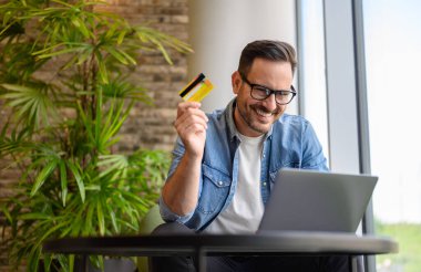 Erkek profesyonel kredi kartı tutuyor ve ofiste otururken dizüstü bilgisayardan sipariş veriyor.