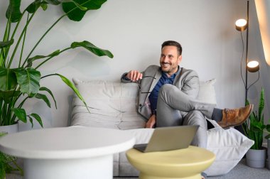 Erkek girişimci, elinde gözlük tutarken ve iş hakkında düşünürken modern ofiste koltukta dinleniyor.