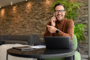 İş yerinde dizüstü bilgisayar kullanırken cep telefonuyla konuşan yakışıklı bir menajerin portresi.