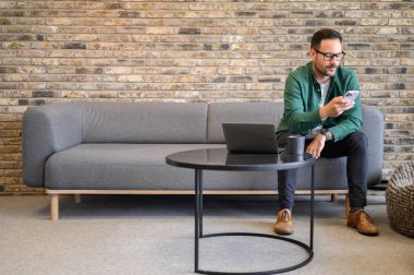 Dizüstü bilgisayarı ve kahve fincanı ile ofisteki koltukta akıllı telefondan mesaj atan kendine güvenen bir serbest yazar.