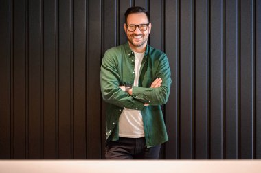 Gözlüklü başarılı erkek müdürün portresi ve kollarını kavuşturup ofiste siyah duvarın önünde duruyor.