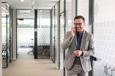 Başarılı erkek girişimci ofiste cam kapının önünde dururken gülümseyip telefonla konuşuyor.