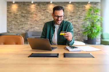 Ofiste otururken kredi kartıyla faturaları ödeyen gülümseyen erkek muhasebeci.