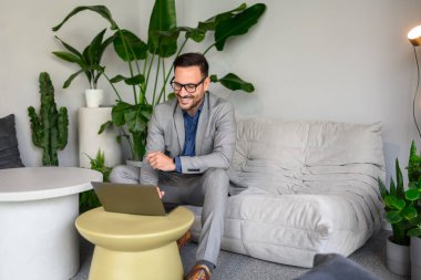 Modern işyerindeki kanepede otururken bilgisayardan e-postaları okuyan başarılı bir erkek yönetici.