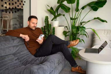 Ofisteki kanepede otururken elinde gözlük tutan ve e-postaları akıllı telefondan okuyan ciddi bir genç.