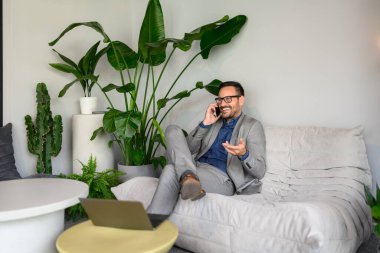 Kendine güvenen satış müdürü iş yerinde rahat bir koltukta otururken telefonla konuşuyor.